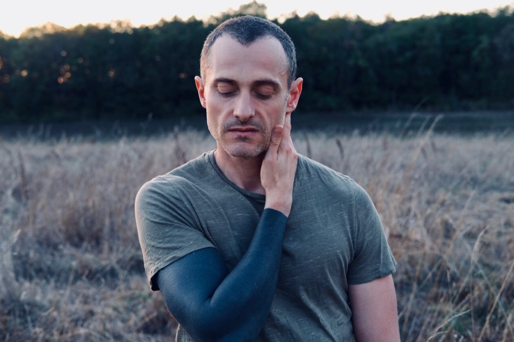 Jean-Baptiste Del Amo headshot of the author looking at the ground, one hand to his neck, displaying a solid black tattoo sleeve.