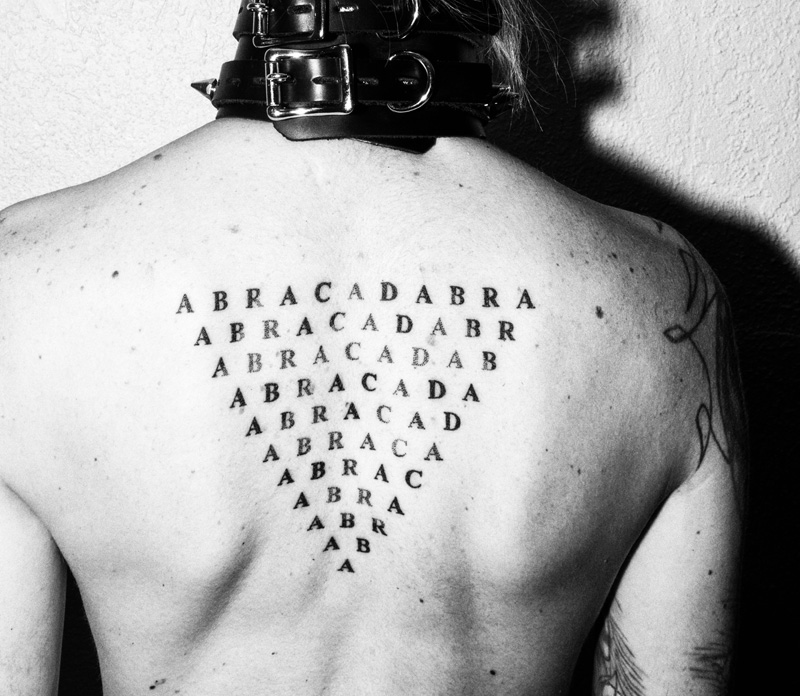 Black and white photo of Rexx's triangular back tattoo, the word ABRACADABRA in serif font repeated with one letter removed until there is only an "A" remaining. Above this, two thick leather collars stacked, the buckle of one lined up on top of the other.