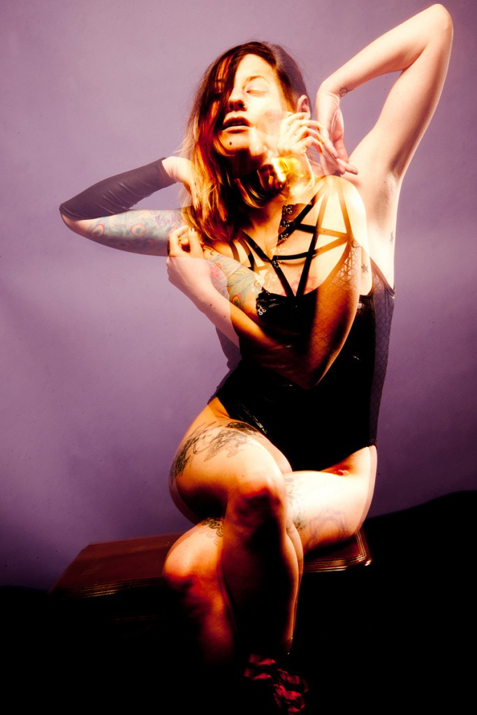 Dreamy double exposure photo of Madison Rexx, a young female alt-model with shoulder length hair and tattoos, seated cross-legged before a purple sheet in two positions: elbows raised on either side as if rising from slumber, and arms embracing her torso. She is wearing a metal choker and a black bodysuit featuring straps in the form of an inverted pentagram over her upper chest.